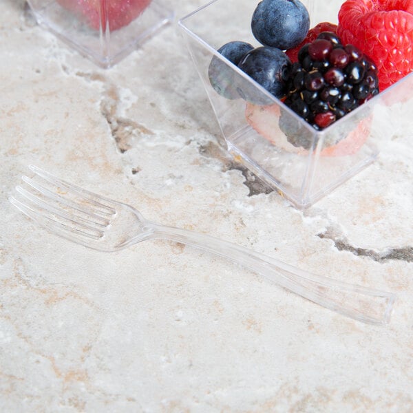 A small clear plastic tasting fork placed on a light-colored stone surface next to a container of mixed berries.