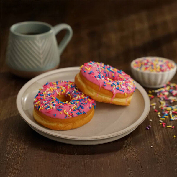 A plate with two pink iced donuts topped with colorful sprinkles, accompanied by a cup of coffee and a small bowl of extra sprinkles.