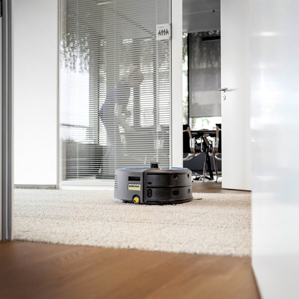 A Karcher Kira CV 50 BP cordless robotic vacuum cleaner on a carpeted floor in an office setting.