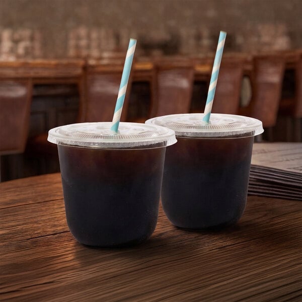 A pair of clear plastic cold cups with flat lids and striped straws, filled with a dark beverage, sitting on a wooden table.