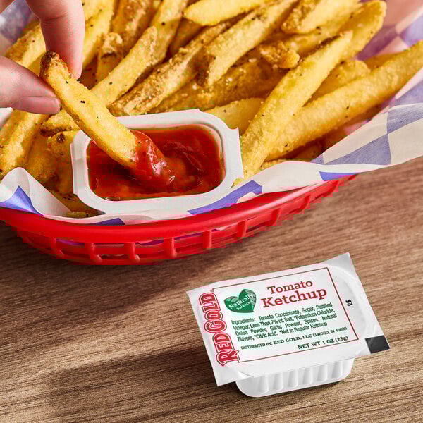 A single-serve cup of Red Gold Naturally Balanced Tomato Ketchup next to a basket of French fries.