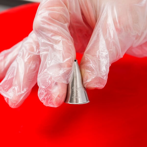 A metal basketweave piping tip held by a gloved hand against a red background.