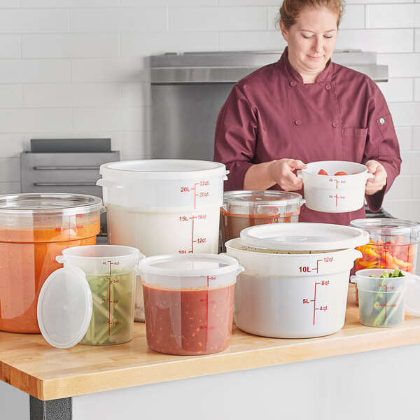 A clear round polycarbonate food storage container with red measurement markings, shown among other containers on a kitchen counter.