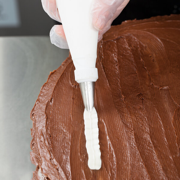 A piping bag fitted with a metal leaf piping tip is being used to decorate a chocolate-frosted cake.