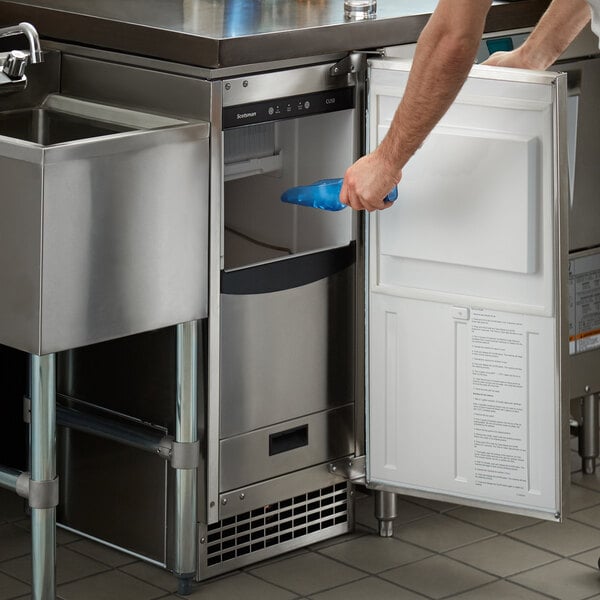 A stainless steel undercounter ice machine with an open door, showing a person using a blue scoop to access the ice bin.