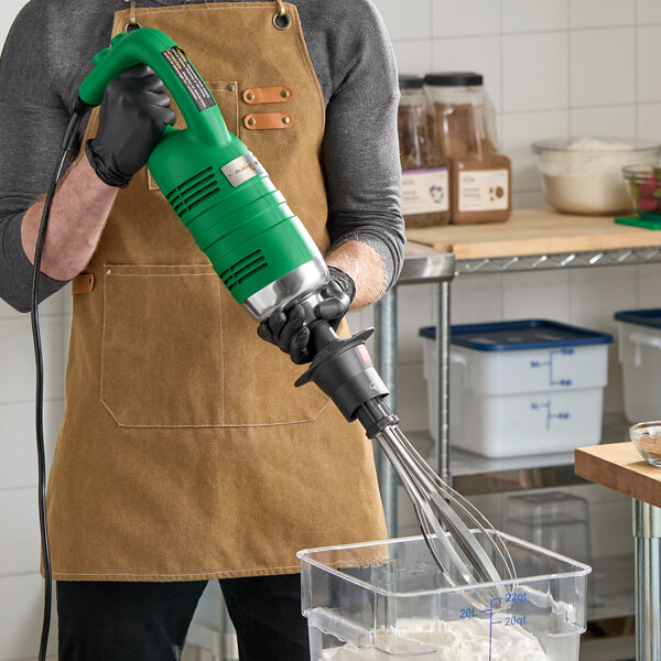 A green heavy-duty immersion blender with a whisk attachment being used in a commercial kitchen.