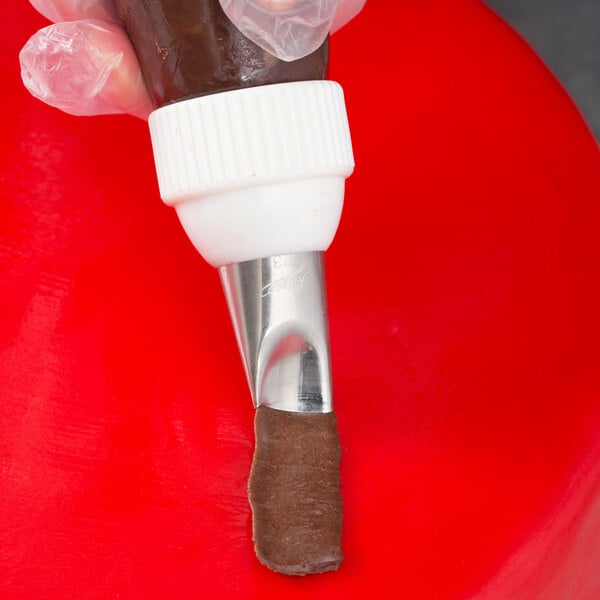 A metal piping tip with a curved petal shape being used to pipe chocolate frosting onto a red surface.