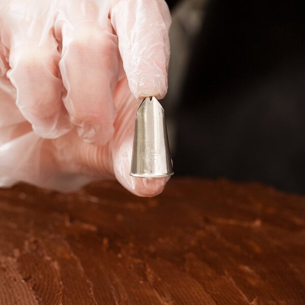 A metal leaf piping tip held by a gloved hand above a chocolate-frosted surface.