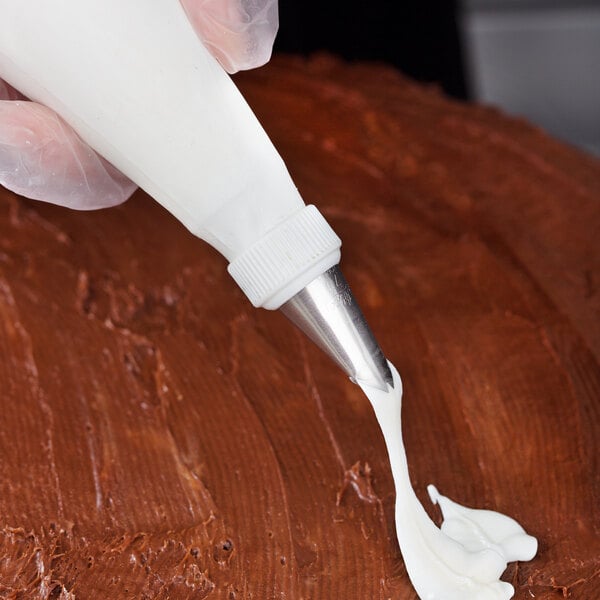 A metal leaf piping tip attached to a piping bag being used to apply white icing onto a chocolate-frosted surface.