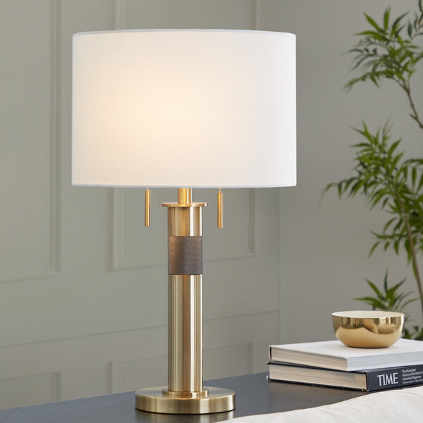 A gold table lamp with a cylindrical base and a white linen drum shade.