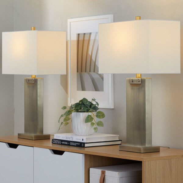 A pair of antique brass table lamps with white linen rectangular shades placed on a wooden surface.