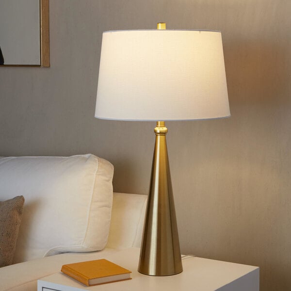 A gold table lamp with a conical base and a white linen drum shade.