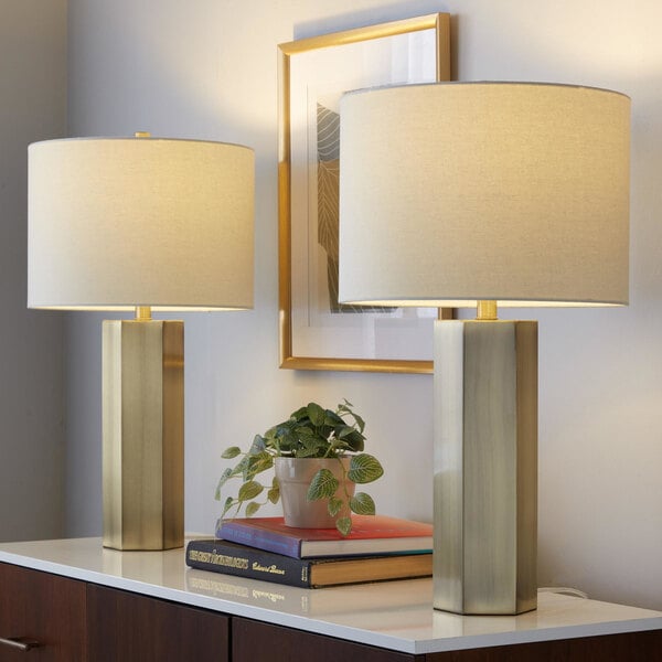 A pair of antique brass table lamps with white linen drum shades placed on a white surface.
