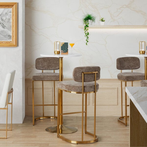 A pair of gold metal barstools with brown fabric backs and seats placed at a high table in a modern setting.