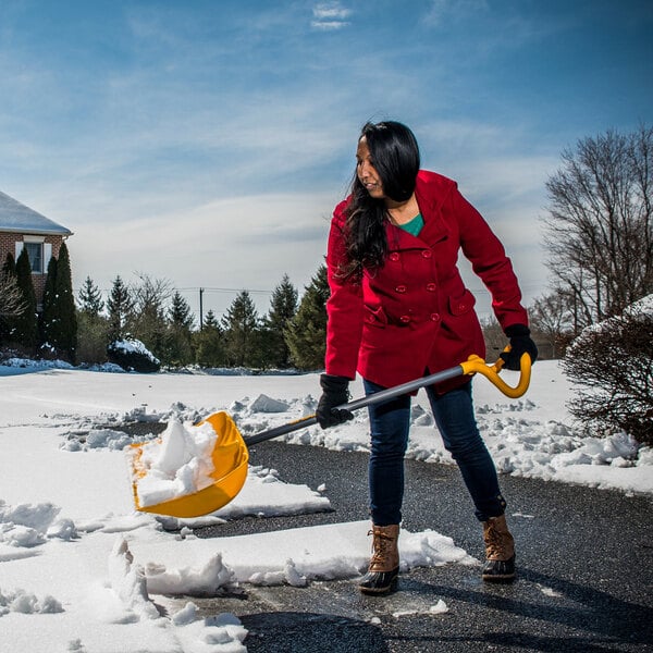 A yellow True Temper 19-inch poly blade combo snow shovel and pusher with a wear strip and VersaGrip handle being used to clear snow from a driveway.