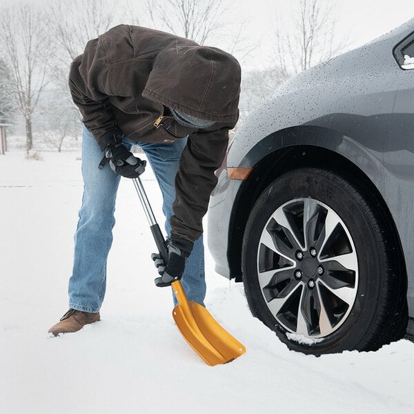 A person using a True Temper 3-in-1 folding emergency snow shovel with an aluminum blade to clear snow near a car tire.