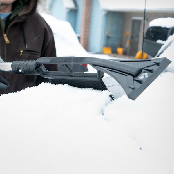 A 3-in-1 folding emergency snow shovel with an aluminum blade, ice scraper, and brush being used to clear snow from a vehicle.