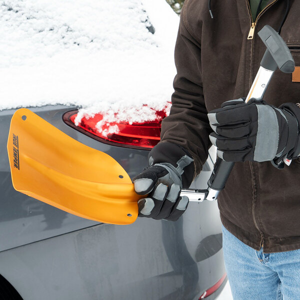 A person holding a True Temper 3-in-1 folding emergency snow shovel with an orange aluminum blade, designed for snow removal and equipped with an ice scraper and brush.