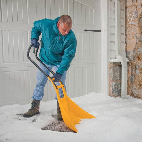 A yellow True Temper SnoBoss 26-inch poly blade snow shovel with a multi-grip handle being used to clear snow from a driveway.