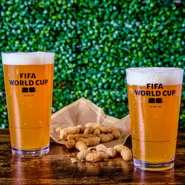 A pair of clear plastic mixing glasses with the FIFA World Cup 2026 logo, filled with beer and placed on a wooden surface next to a pile of peanuts.