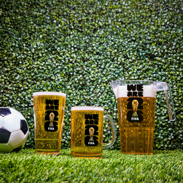 A set of FIFA World Cup 2026 branded plastic beer mugs and a pitcher filled with beer, displayed on artificial grass next to a soccer ball.