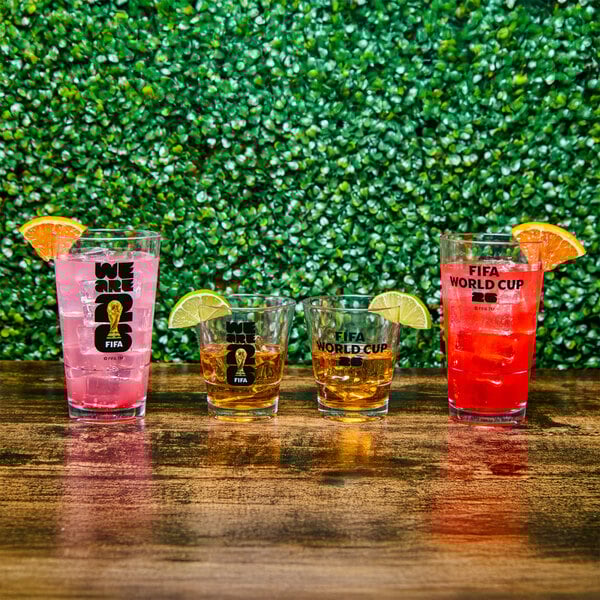 A set of FIFA World Cup 2026 themed plastic pint and rocks glasses with black logos, each filled with colorful beverages and garnished with citrus slices.