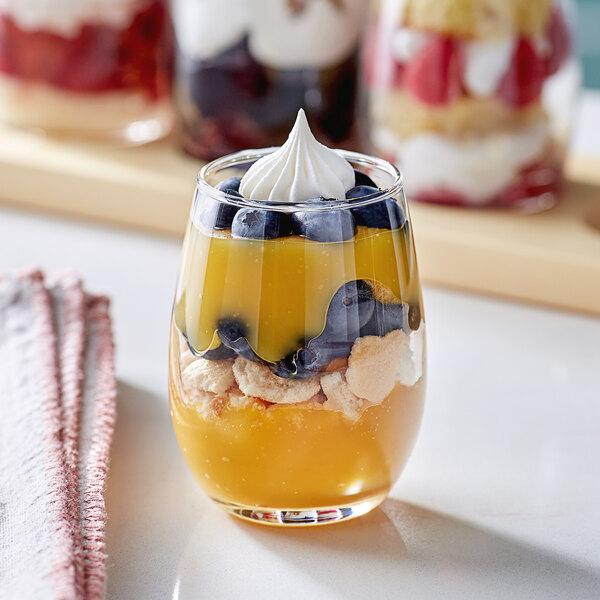 A small stemless wine tasting glass filled with layered dessert, including blueberries, yellow custard, crumbled cookies, and a dollop of whipped cream on top.