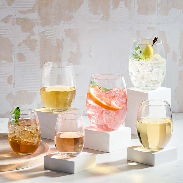 A set of six clear, stemless wine glasses filled with various beverages, displayed on white blocks.