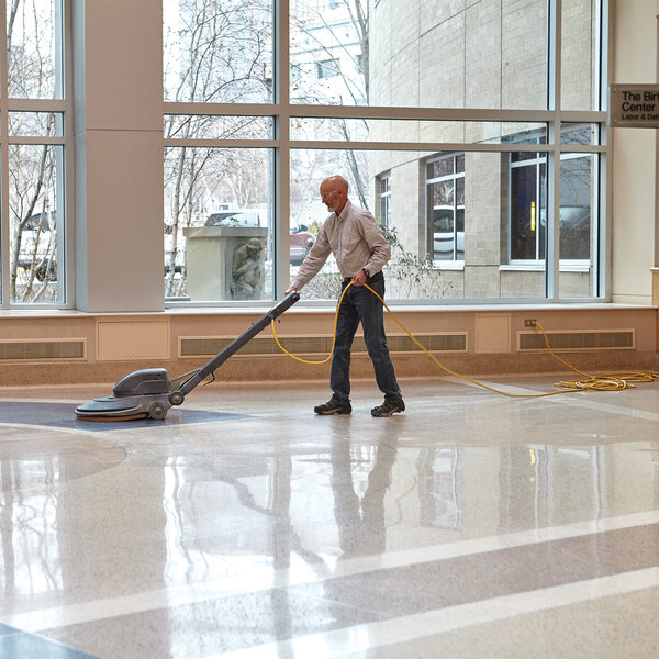 A person using a floor burnishing machine with a tan burnishing pad on a large, shiny indoor floor.