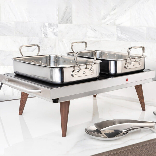 A stainless steel warming shelf with two rectangular pans and wooden legs, designed for keeping food warm.