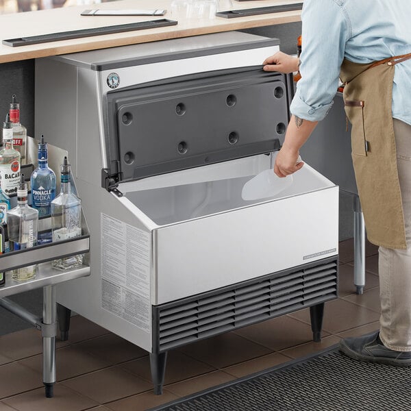 A commercial undercounter ice machine with a front-opening bin, shown in use in a bar setting.
