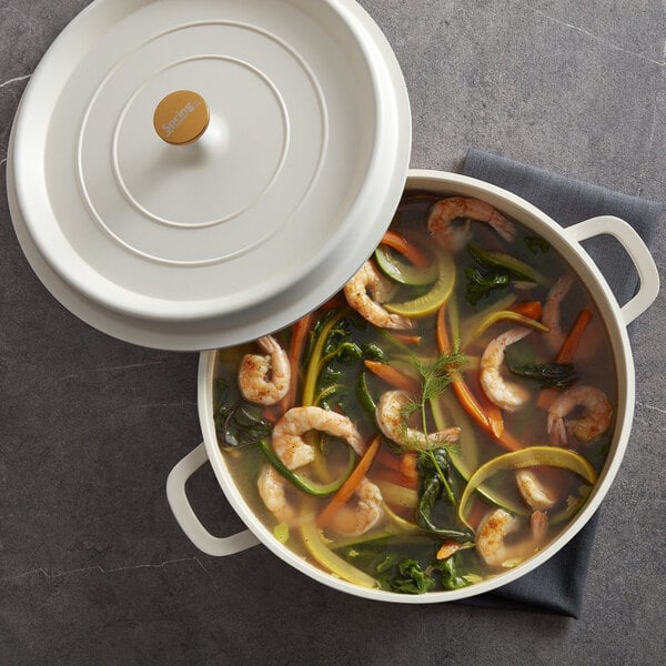 A white round casserole dish with handles and a lid, filled with shrimp and vegetable soup.