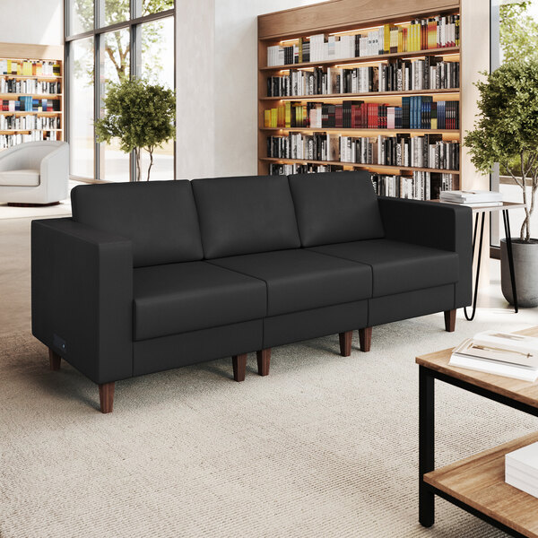 A black three-seat sofa with wooden legs and built-in charging banks, placed in a modern library setting.