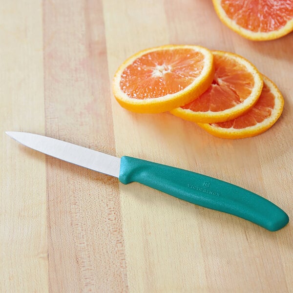 A small paring knife with a kale green handle and a stainless steel blade, shown next to sliced oranges on a wooden surface.