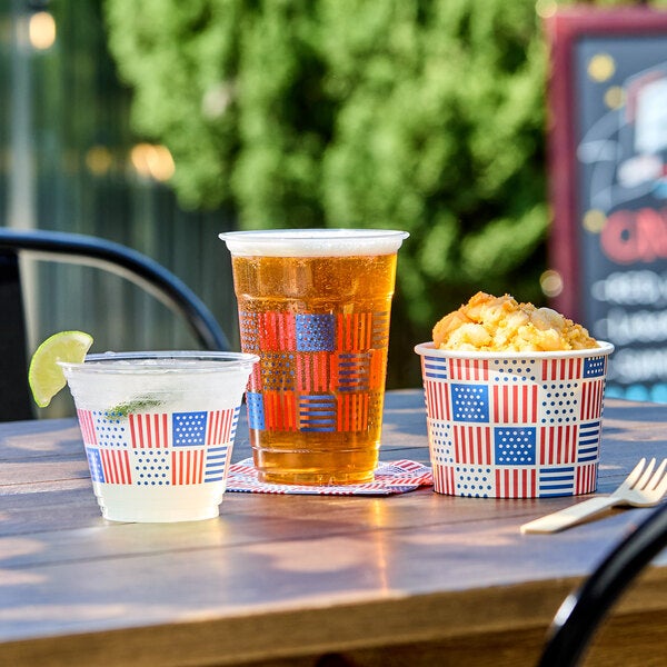 A clear plastic squat cold cup with a patriotic American flag print, filled with a beverage and garnished with a lime wedge.