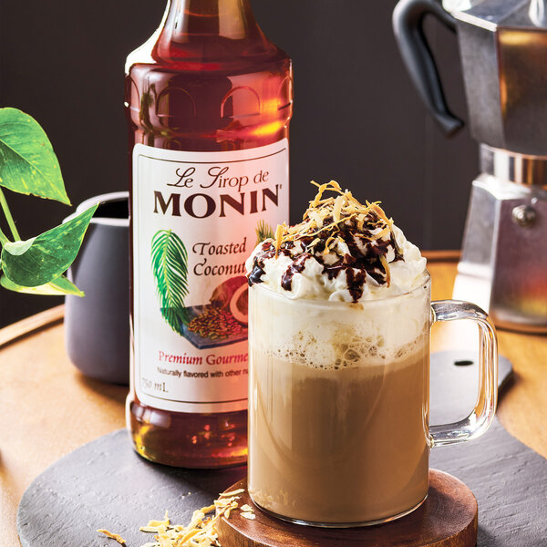A bottle of Monin Premium Toasted Coconut Flavoring Syrup next to a glass mug of a whipped cream-topped beverage garnished with toasted coconut and chocolate drizzle.