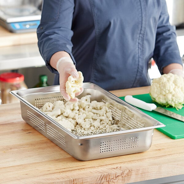 Cauliflower being cut on a green cutting board and placed in a Vollrath stainless steel pan with a hole in the bottom.