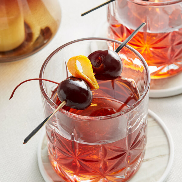 A black bamboo twisted food pick skewering two cherries and an orange peel, placed in a glass of cocktail.