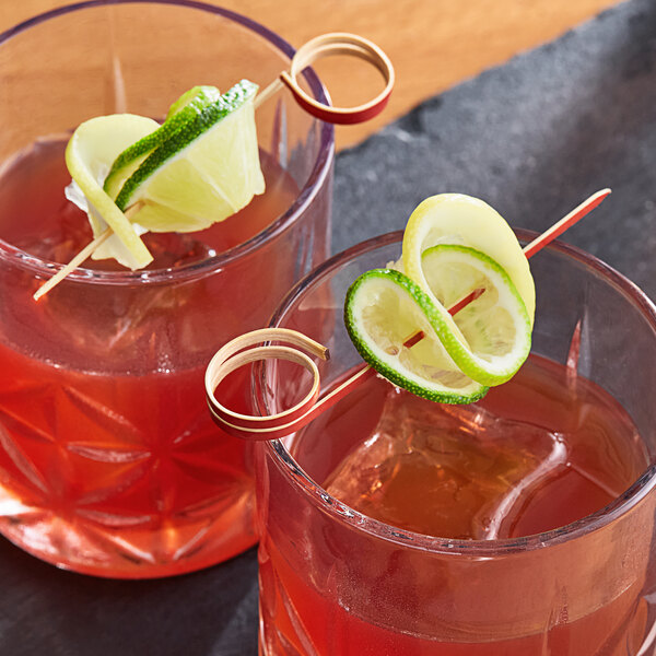 A pair of red bamboo loop food picks skewering lime garnishes in two glasses of red beverages.
