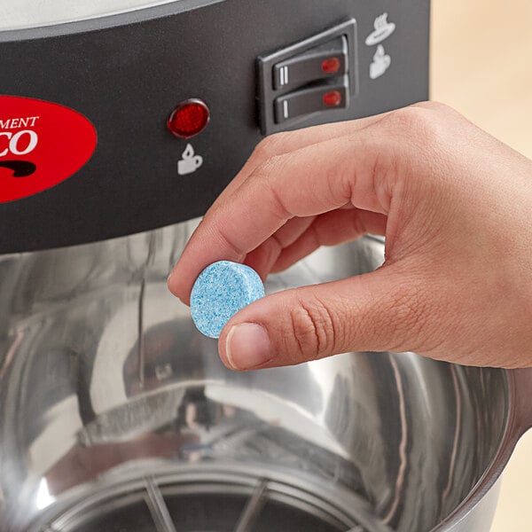 A hand holding a blue and white coffee equipment cleaning tablet above a metal coffee machine.