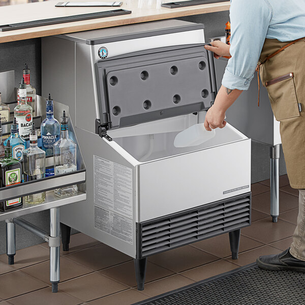 A stainless steel undercounter ice machine with a built-in storage bin, shown with its door open in a commercial bar setting.
