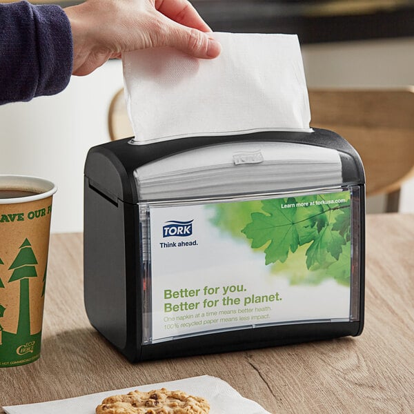 A black tabletop interfold napkin dispenser with a clear front panel displaying a green and white label.