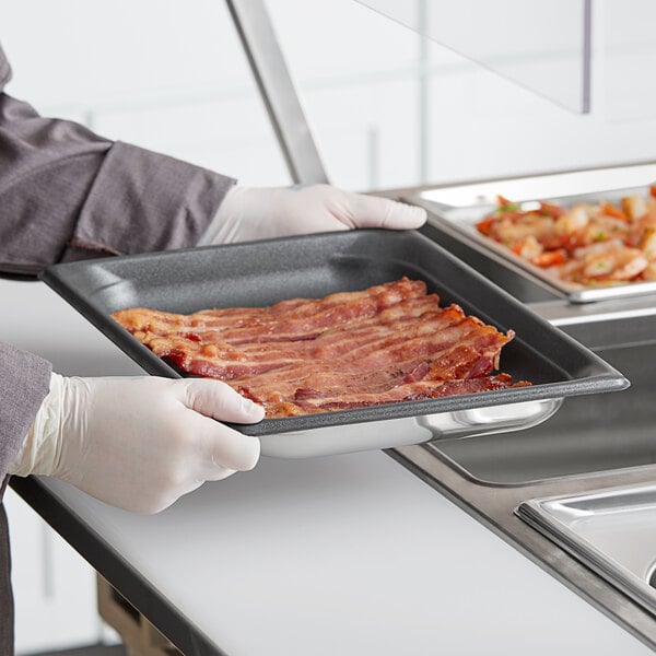A person in a white coat holding a Vollrath stainless steel steam table pan filled with bacon.