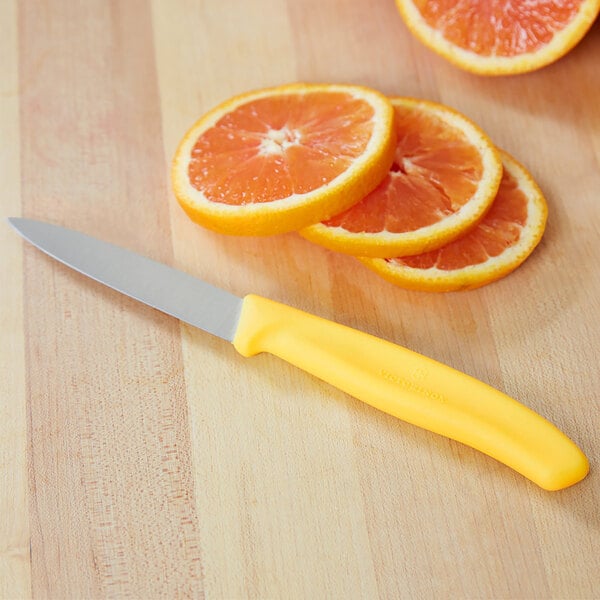 A small paring knife with a lemon yellow handle next to several slices of orange on a wooden surface.
