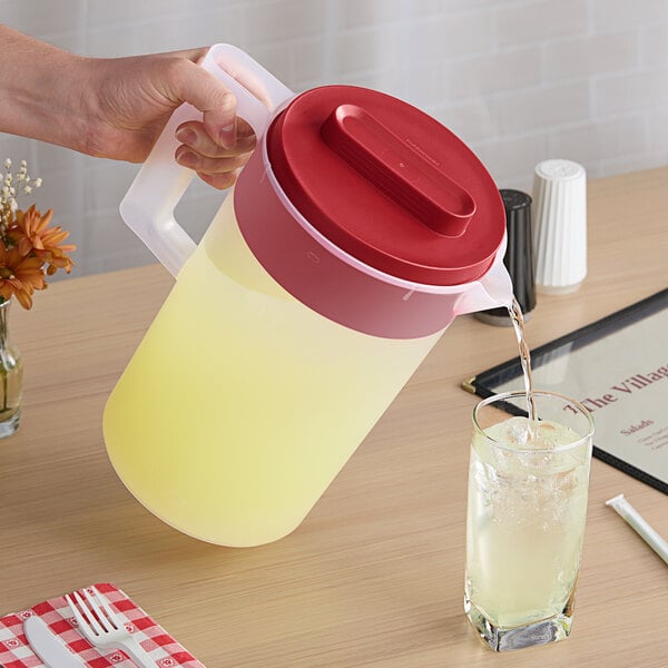A large plastic pitcher with a red lid being used to pour a yellow beverage into a glass.