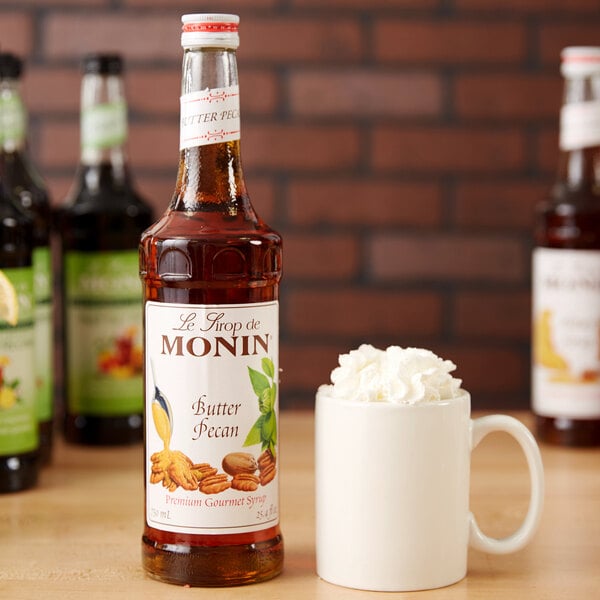 A bottle of Monin Premium Butter Pecan Flavoring Syrup next to a white mug topped with whipped cream.