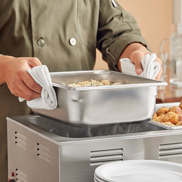 A stainless steel 1/2 size, 4-inch deep steam table or hotel pan being handled by a person using towels.