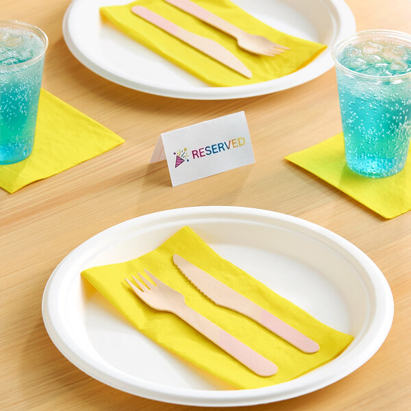A table setting with white plates, yellow napkins, wooden cutlery, blue drinks in clear cups, and a 'Reserved' sign.