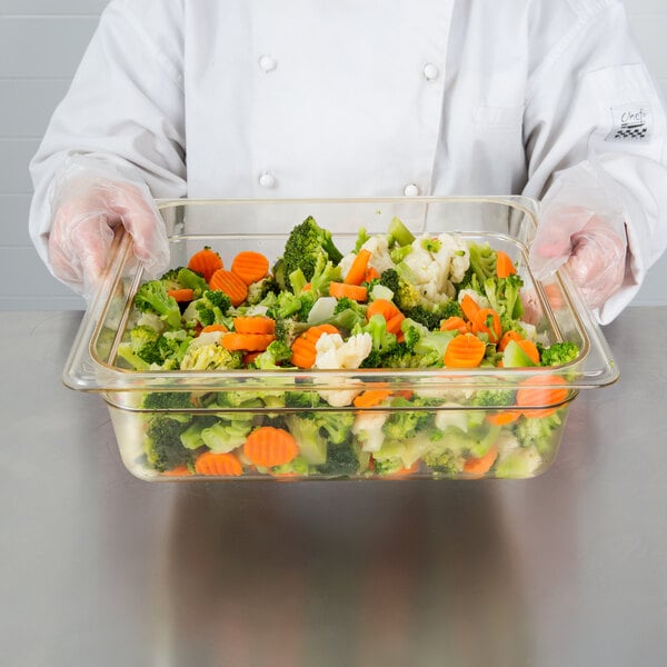 A clear amber high heat plastic food pan filled with mixed vegetables, held by a person wearing a white chef coat and gloves.