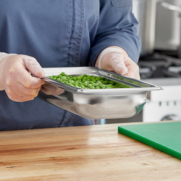 A person in a chef's uniform holding a Vollrath stainless steel hotel pan of green peas.